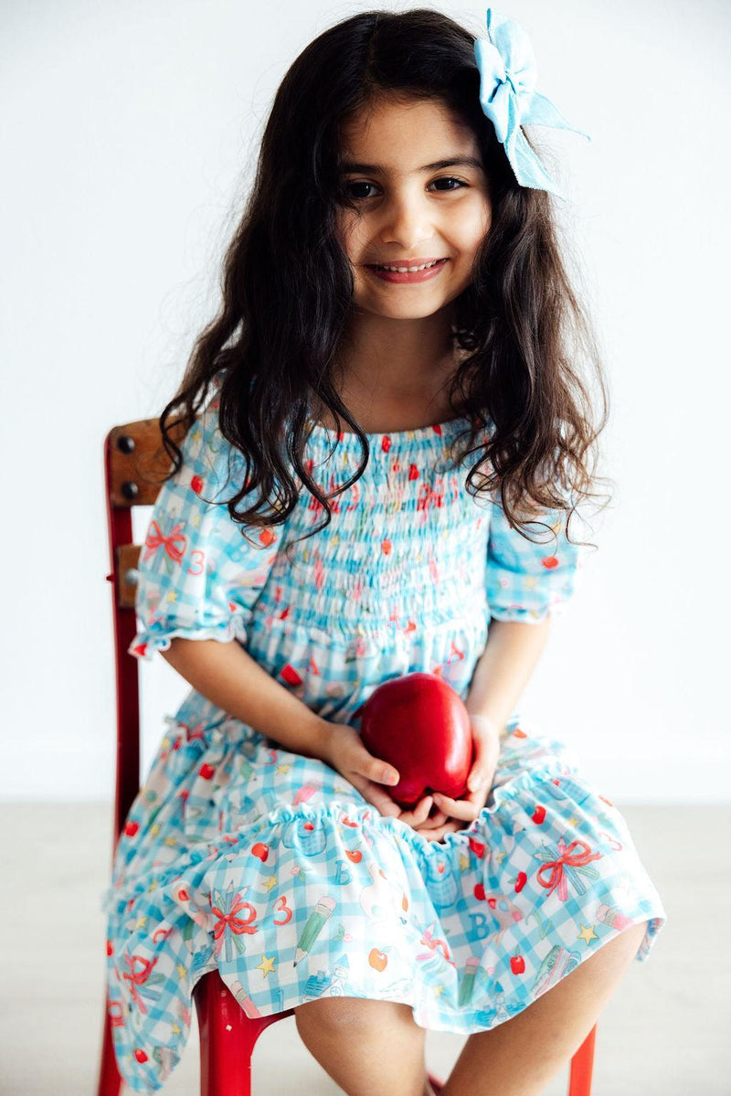 A joyful girl in a blue Honor Roll Smocked Ruffle Dress by Mila & Rose®, sitting on a red chair with an apple in hand, exuding style and confidence.