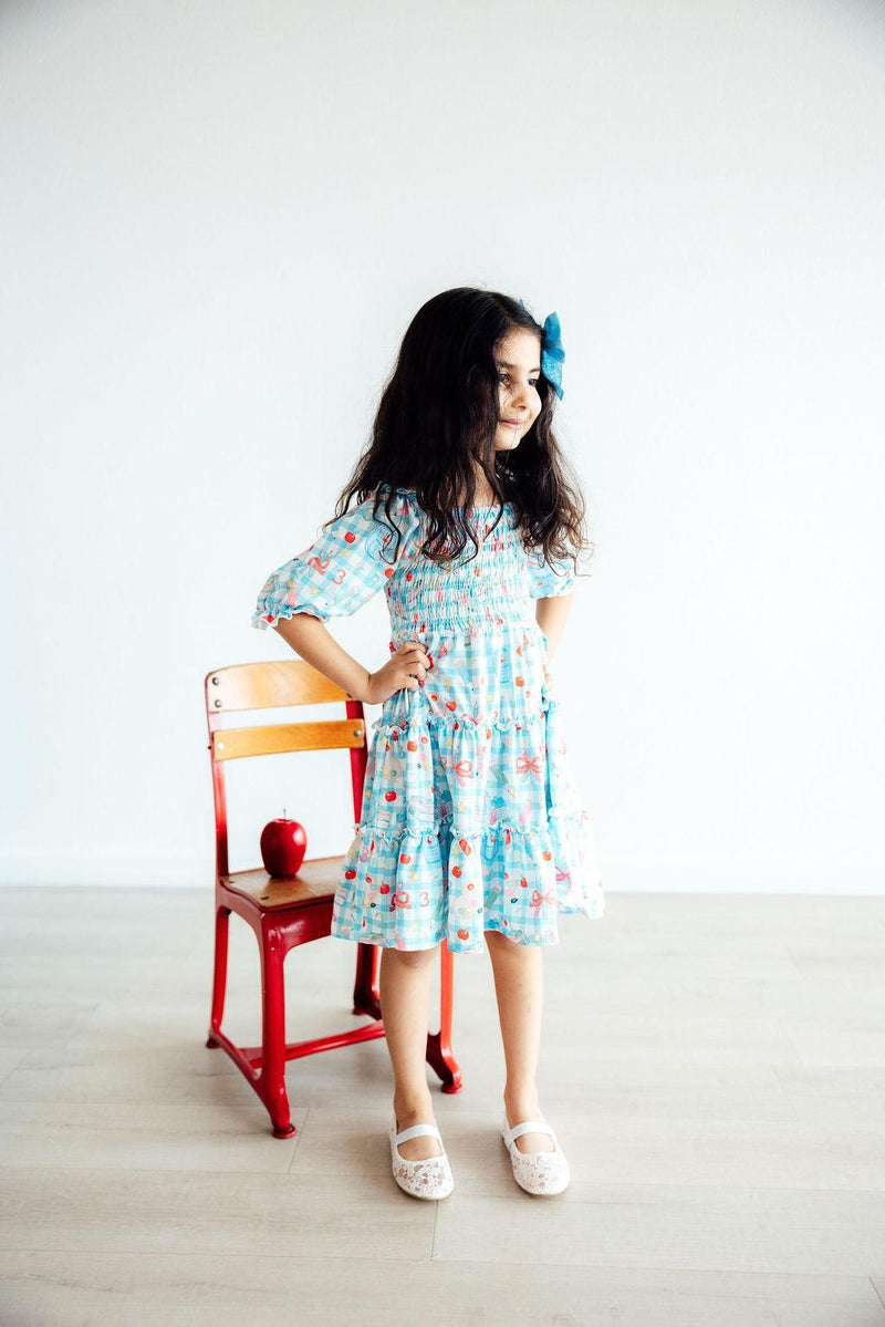 A confident girl in the blue Honor Roll Smocked Ruffle Dress by Mila & Rose®, striking a pose beside a red chair with an apple, ready to take on the day.