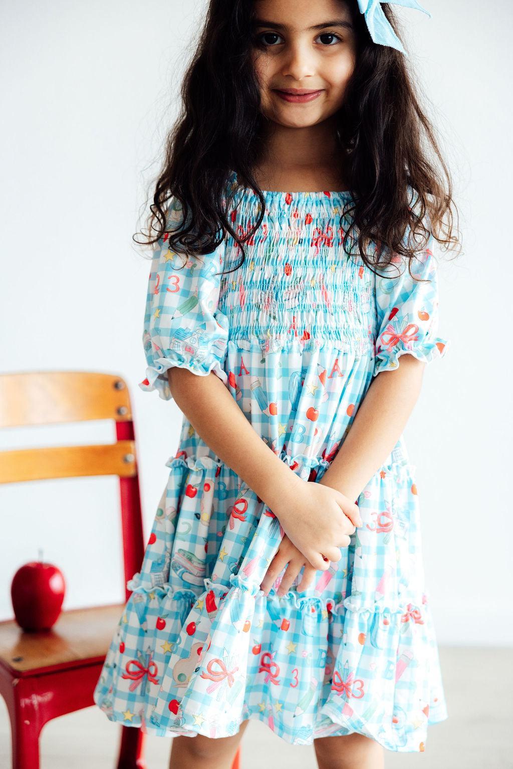 A young girl wearing the Honor Roll Smocked Ruffle Dress by Mila & Rose®, featuring a colorful design with playful patterns and a twirl-worthy ruffle skirt, perfect for any little fashionista!