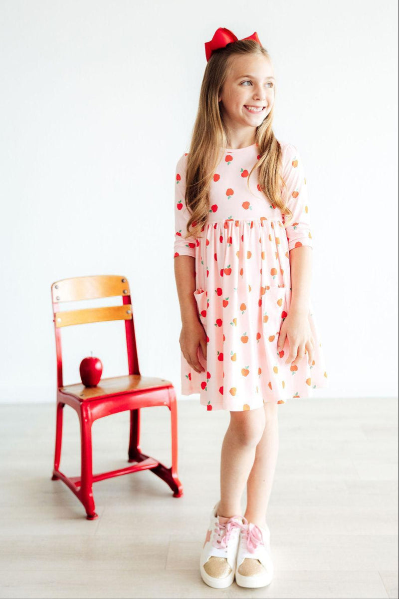 A joyful girl smiling in an 'Apple a Day' 3/4 Sleeve Pocket Girls Twirl Dress, standing next to a red chair and an apple, perfect for school or parties.