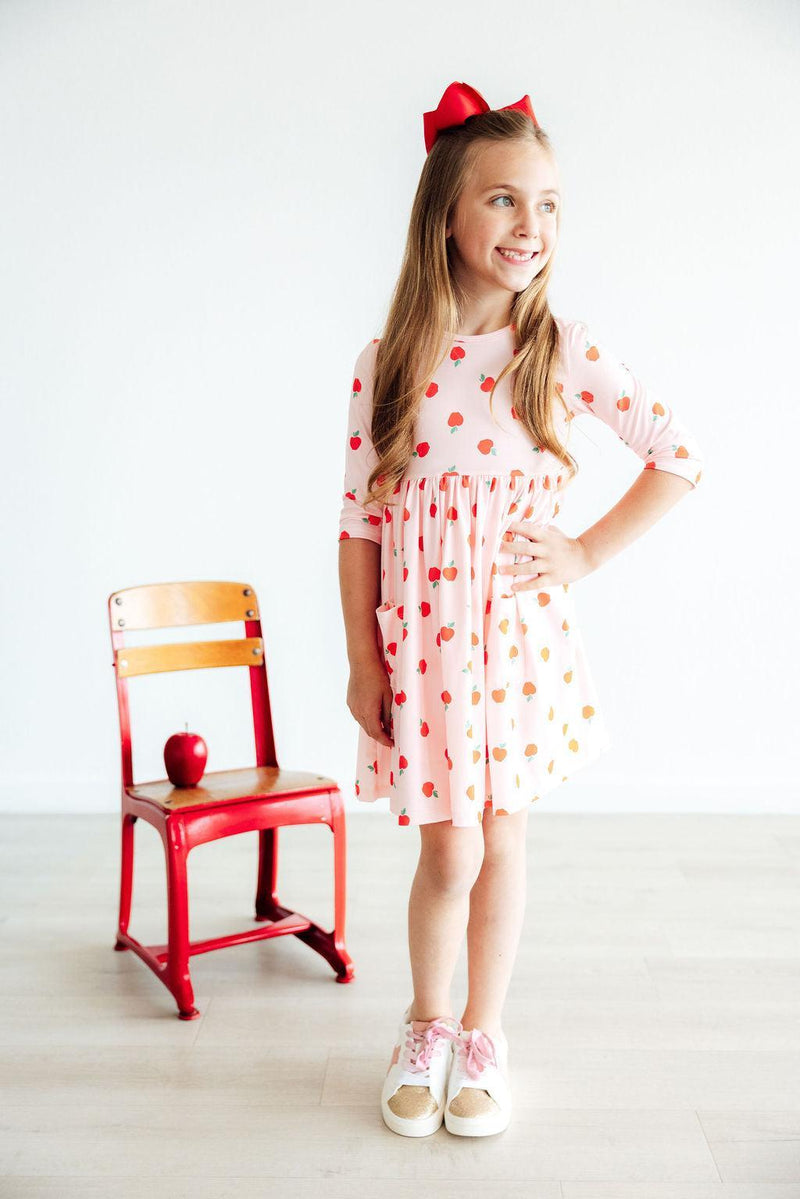 A girl in an 'Apple a Day' 3/4 Sleeve Pocket Girls Twirl Dress smiling while standing beside a red chair with an apple, showcasing a cute everyday outfit.