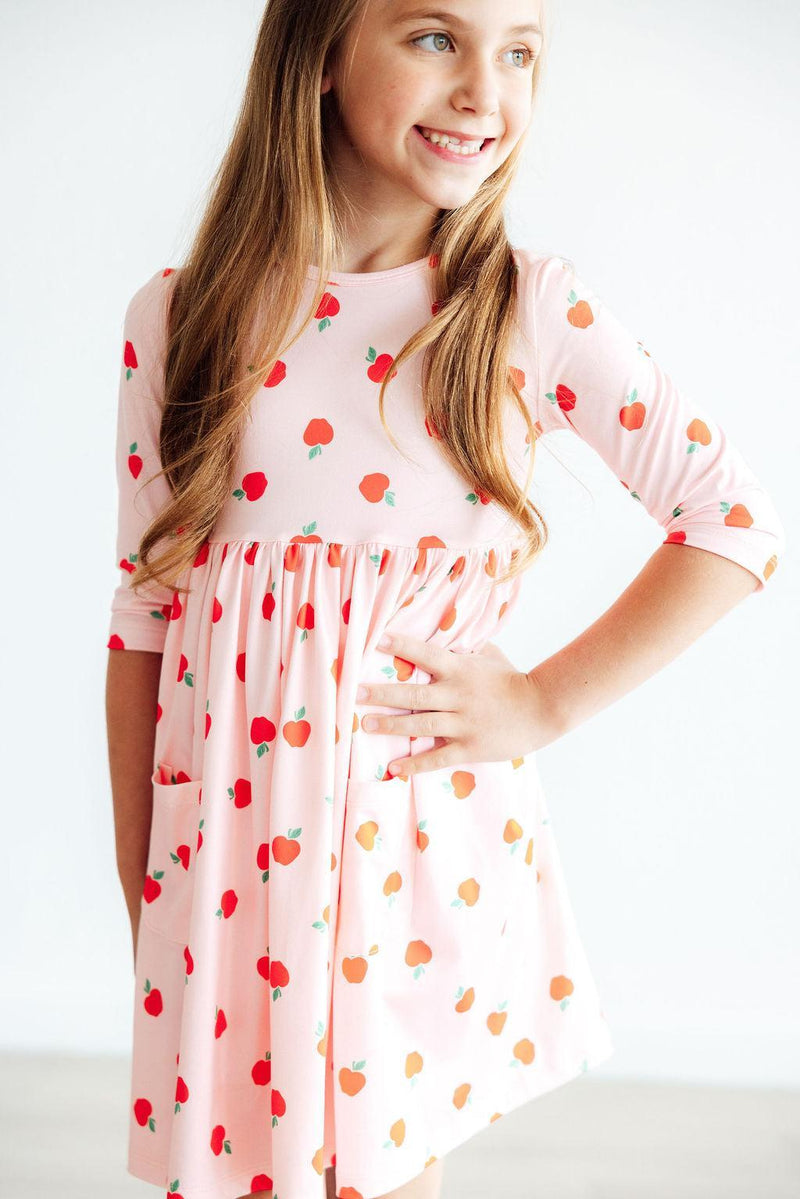 A happy girl posing in an 'Apple a Day' 3/4 Sleeve Pocket Girls Twirl Dress, highlighting its comfy fit and playful design with a pocket.