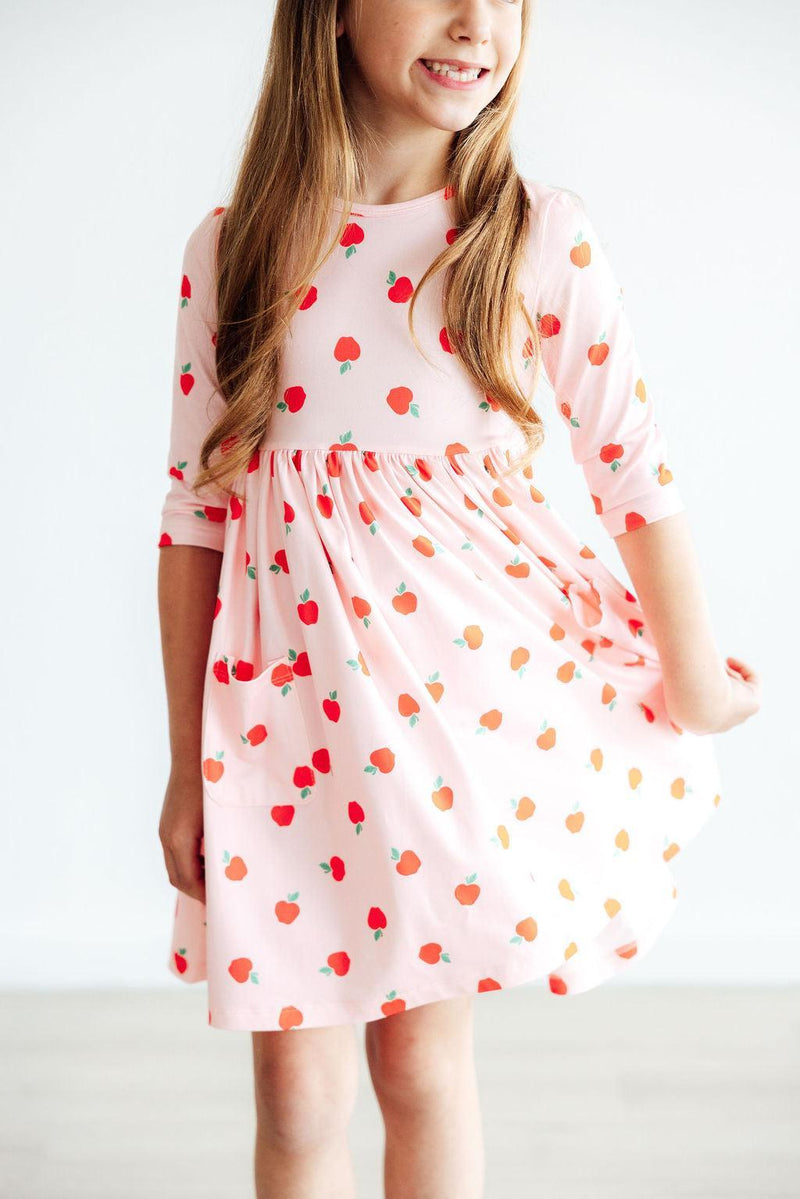 A cheerful girl twirling in an 'Apple a Day' 3/4 Sleeve Pocket Girls Twirl Dress with an apple print, bringing joy and playfulness to her outfit.