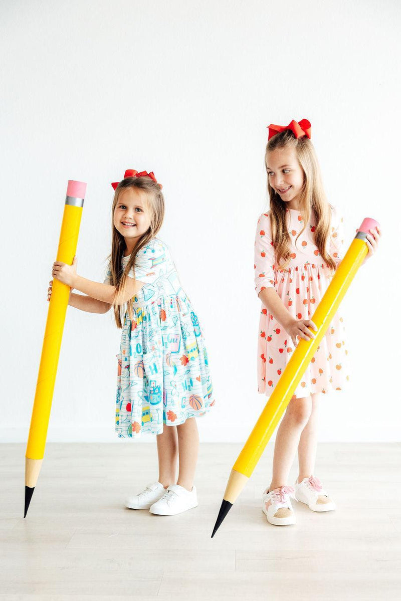 Two girls having fun in 'Apple a Day' 3/4 Sleeve Pocket Girls Twirl Dresses, one in a colorful dress and the other in apple print, holding oversized pencils.