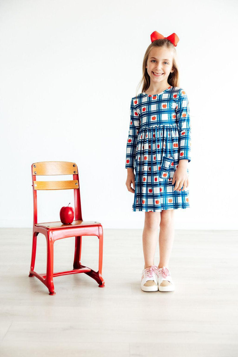 Charming young girl wearing the Plaid Apples L/S Pocket Twirl Dress by Mila & Rose, posing beside a bright red chair, showcasing the playful apple pattern and vibrant blue plaid design, perfect for twirls and fun!