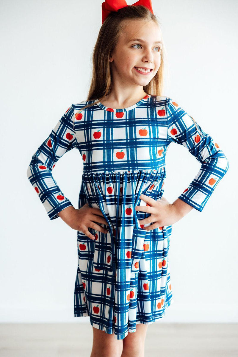 Delightful close-up of a girl in the Plaid Apples L/S Pocket Twirl Dress by Mila & Rose, striking a pose with hands on her hips, highlighting the adorable apple prints and the twirly skirt for stylish playtime!