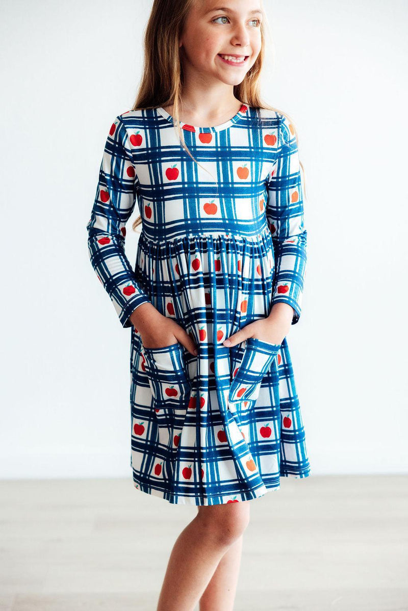 Close-up of a girl confidently modeling the Plaid Apples L/S Pocket Twirl Dress, featuring pockets and a playful apple print for the ultimate twirl.