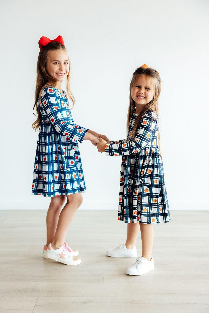 Two girls holding hands, showcasing the adorable Plaid Apples L/S Pocket Twirl Dress with vibrant patterns, perfect for twirling and giggling.