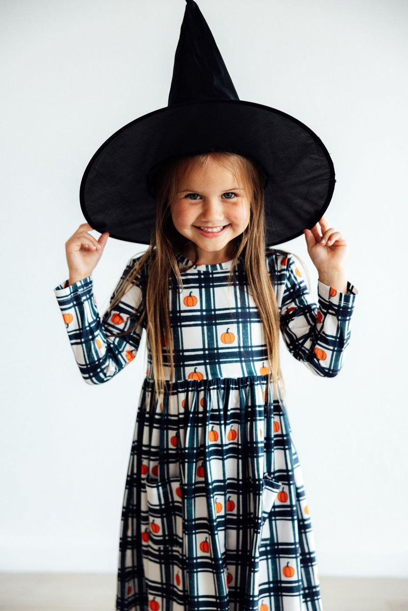 A beaming girl in the Plaid Pumpkins Long Sleeve Pocket Twirl Dress, playfully wearing a black witch hat to celebrate the season.