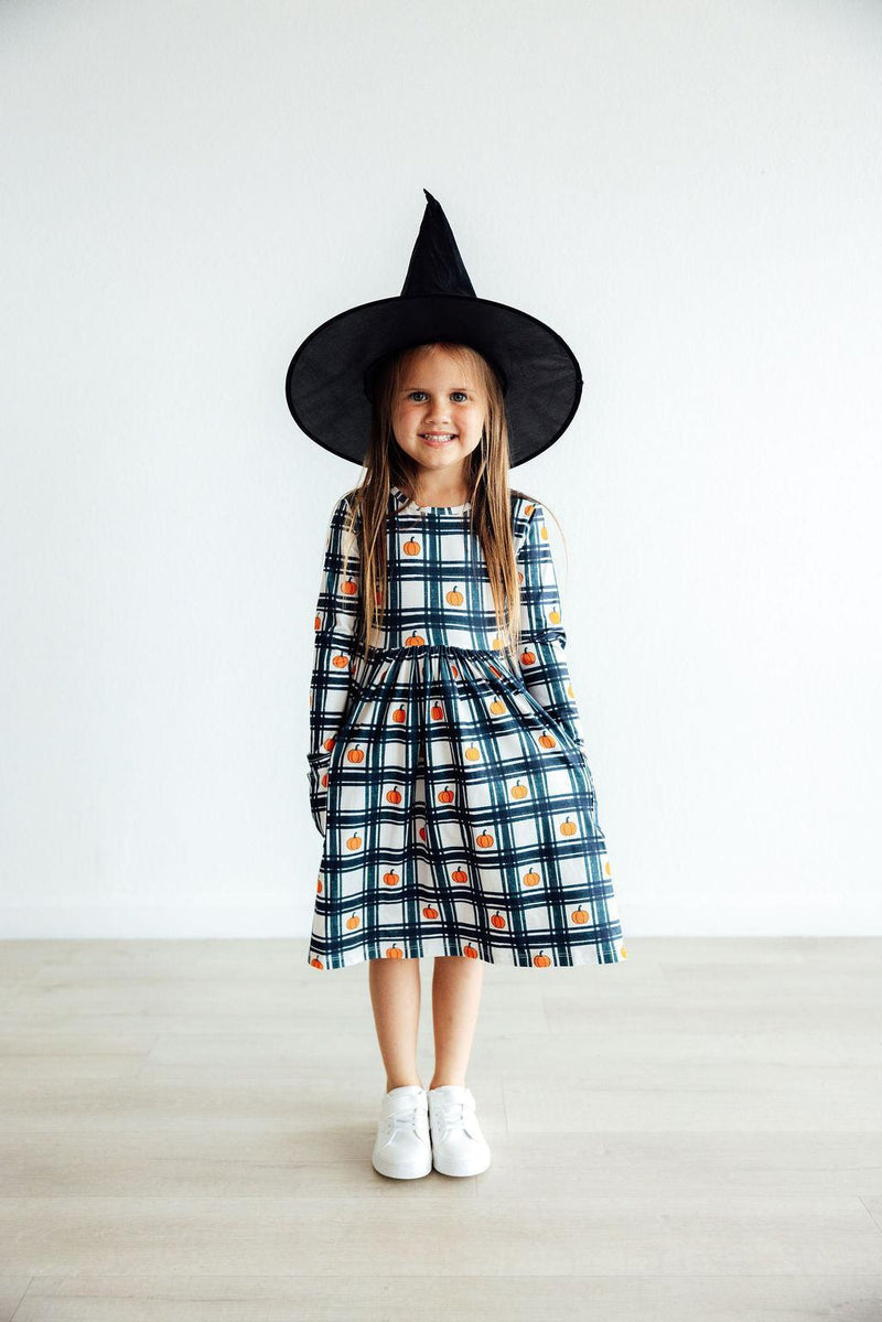 A charming girl posing in the Plaid Pumpkins Long Sleeve Pocket Twirl Dress with a witch hat, ready for autumn adventures.