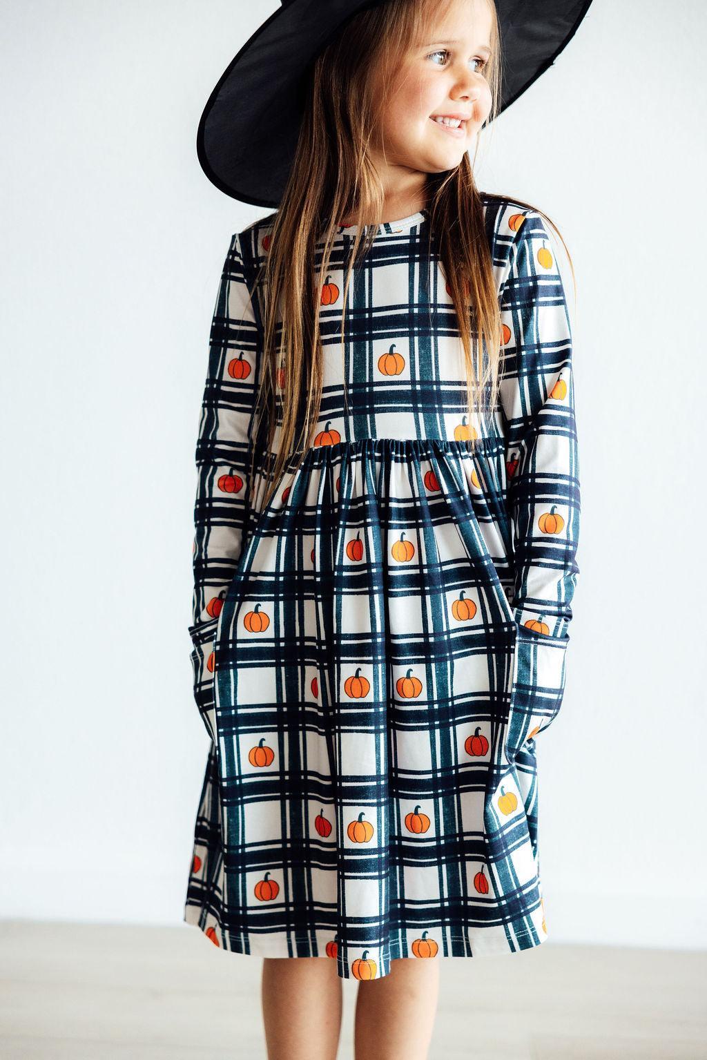 A joyful girl wearing the Plaid Pumpkins Long Sleeve Pocket Twirl Dress with a stylish black hat, showcasing the dress's playful plaid and pumpkin design.