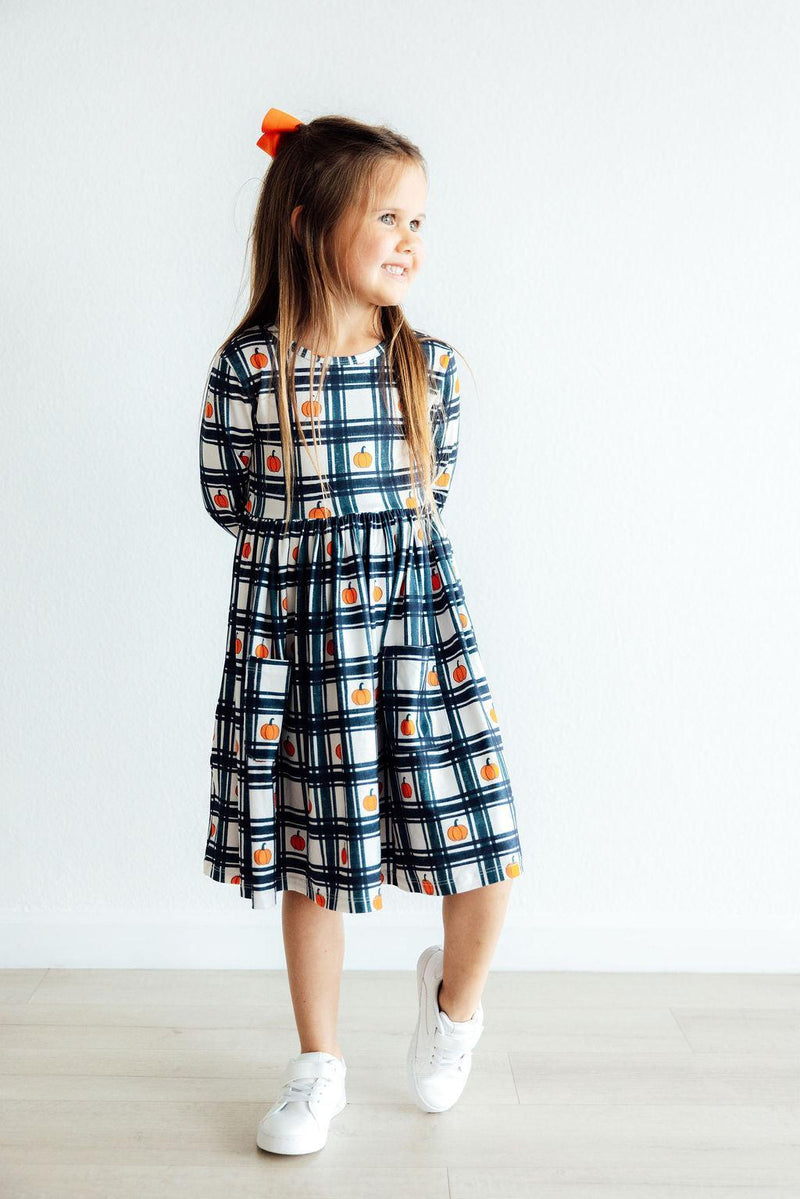 A smiling girl in the Plaid Pumpkins Long Sleeve Pocket Twirl Dress, engaging with a playful look, perfect for twirls and autumn festivities.