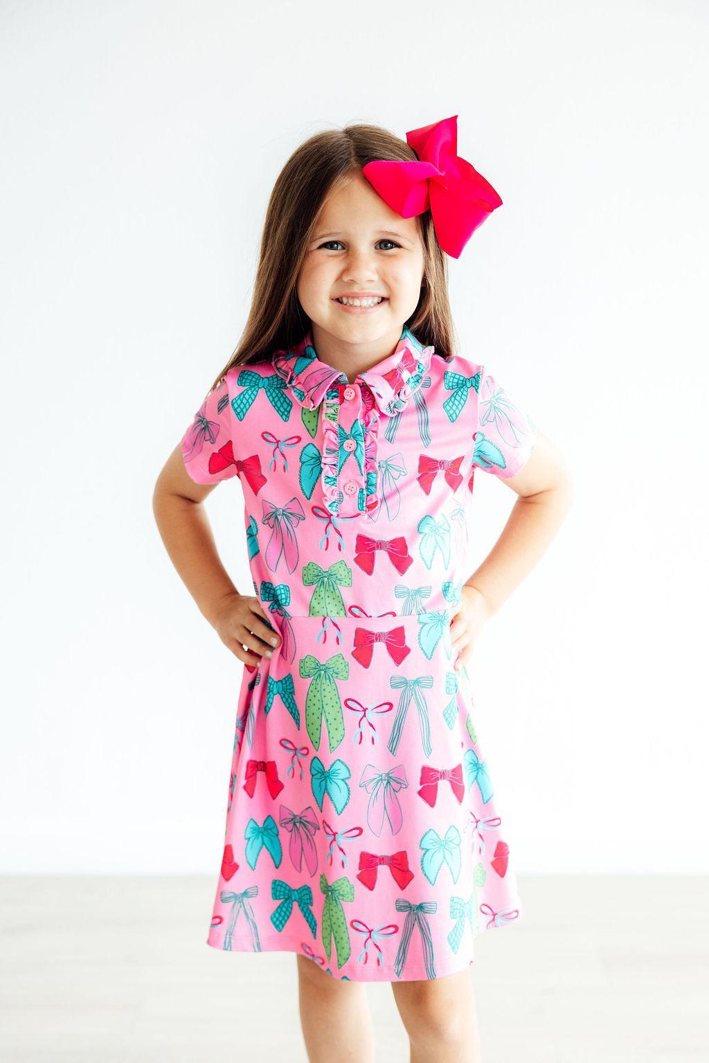 A happy girl wearing the 'Twirls & Ties' Short Sleeve Pink Polo Dress for Girls, featuring colorful bows and leaves in a playful pattern. This stylish pink polo dress for girls is perfect for outdoor fun.
