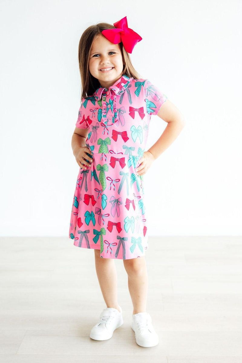 A smiling girl posing confidently in the 'Twirls & Ties' Short Sleeve Pink Polo Dress for Girls, emphasizing the fun design and comfortable fit. This adorable pink polo dress for girls is great for any occasion.