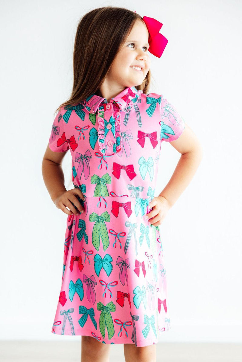 A cheerful girl with a pink bow in her hair, wearing the 'Twirls & Ties' Short Sleeve Pink Polo Dress for Girls. This eye-catching pink polo dress for girls is perfect for twirling and stylish play.