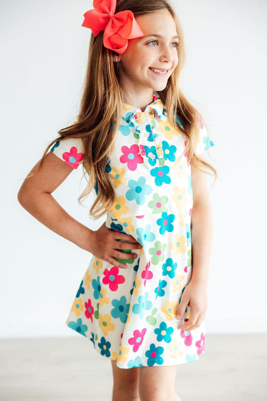 A smiling girl wearing the 'Groovy Blooms' Colorful Short Sleeve Girls Polo Dress, showcasing vibrant floral patterns, complete with a bow in her hair. Perfect girls polo dress for playful occasions.