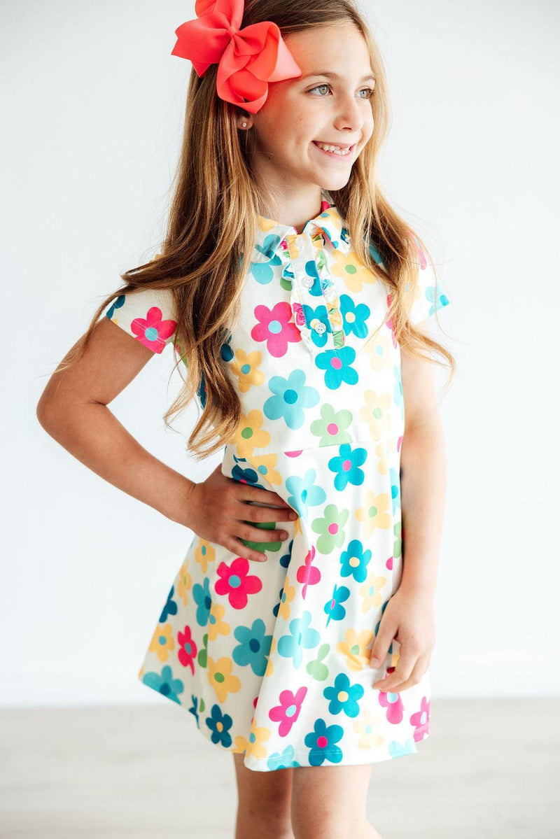A smiling girl wearing the 'Groovy Blooms' Colorful Short Sleeve Girls Polo Dress, showcasing vibrant floral patterns, complete with a bow in her hair. Perfect girls polo dress for playful occasions.