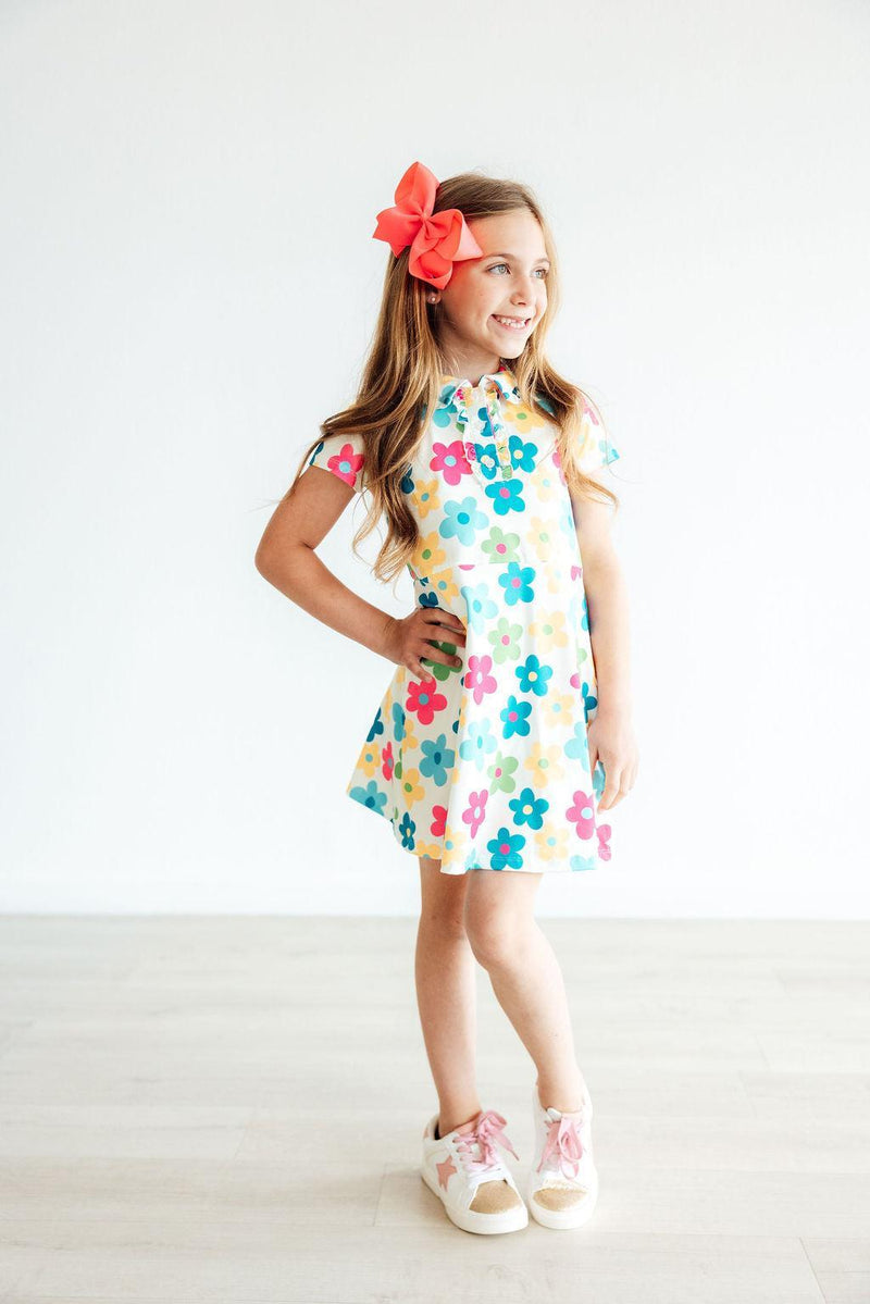 A cheerful girl in the 'Groovy Blooms' Colorful Short Sleeve Girls Polo Dress, adorned with colorful flowers and styled with a hair bow, ideal for a fun day out. Perfect girls polo dress for little fashionistas.