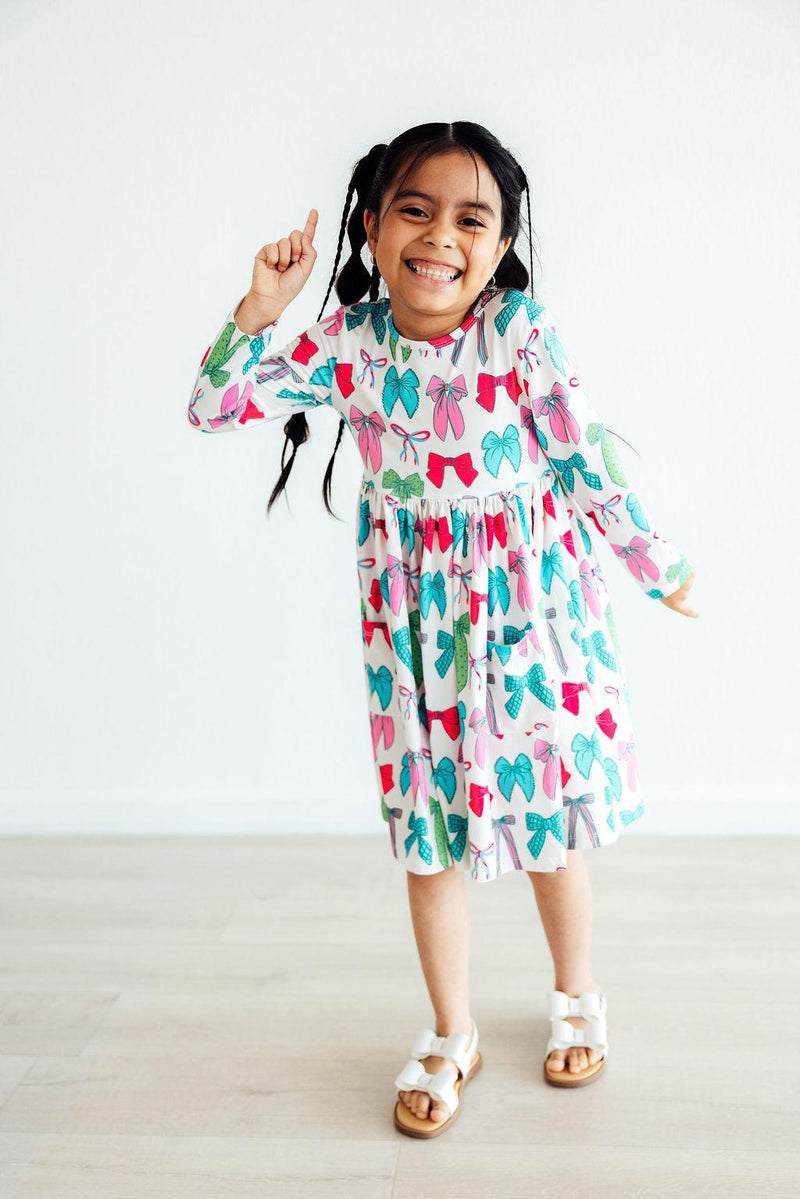 Joyful girl dancing in the Twirls & Ties L/S Pocket Twirl Dress featuring vibrant bow designs, perfect for twirling and fun activities.