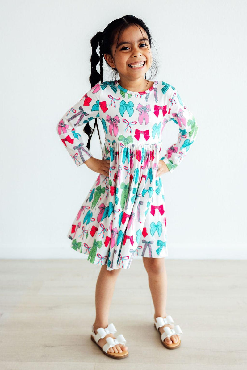 Smiling girl posing confidently in the Twirls & Ties L/S Pocket Twirl Dress, adorned with bright bow patterns, ideal for any playful occasion.