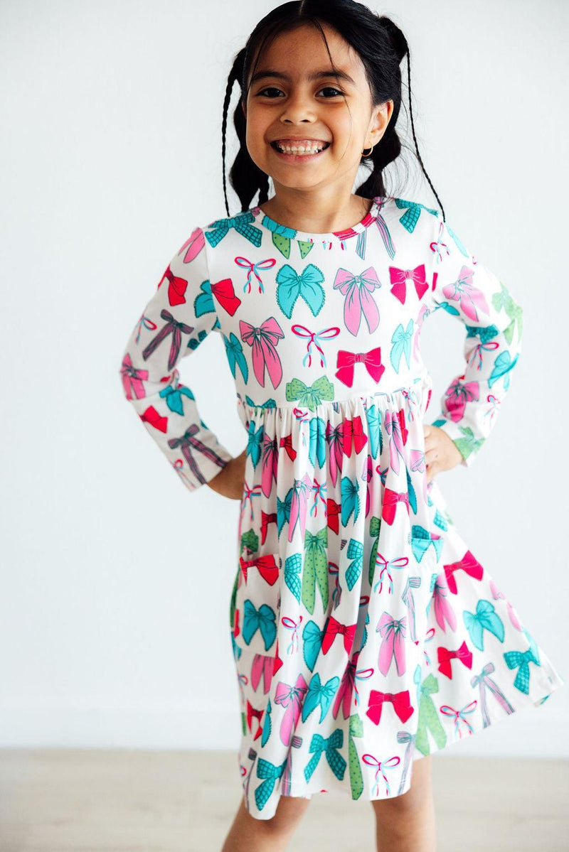 Smiling girl wearing the Twirls & Ties L/S Pocket Twirl Dress by Mila & Rose, featuring playful bow and leaf patterns, perfect for twirling with comfort.