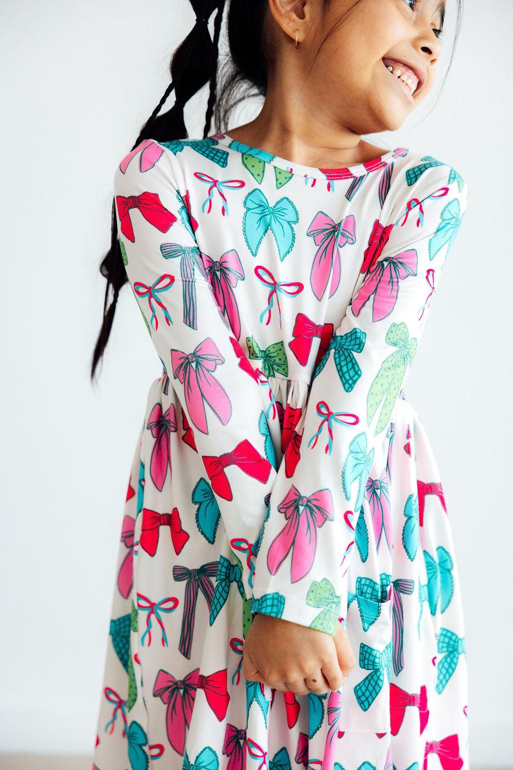 Cute girl happily wearing the Twirls & Ties L/S Pocket Twirl Dress with colorful bow prints, showcasing her playful personality.