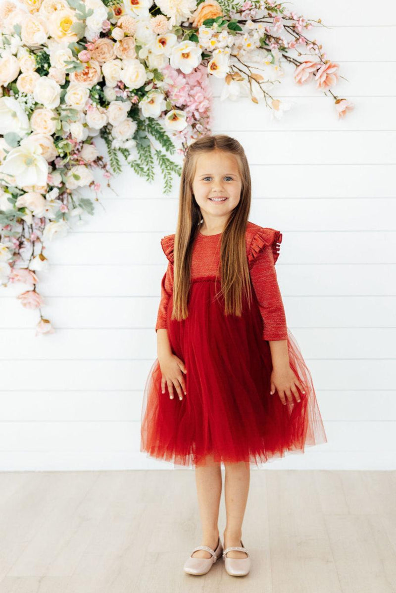 Cute girl smiling in a Pumpkin Spice Shimmer 3/4 Sleeve Tutu Dress standing in front of a floral backdrop.