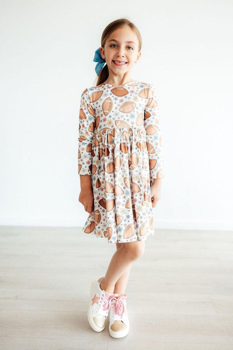 Happy girl wearing the Football Friday Long Sleeve Pocket Twirl Dress with football designs, showcasing a full-circle twirl skirt and stylish sneakers.