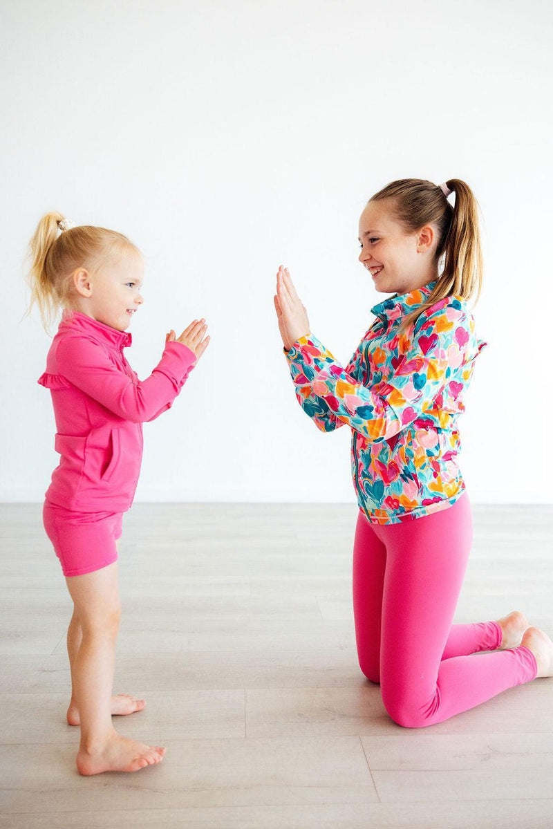 A delighted girl kneeling next to a younger girl, both wearing vibrant active outfits including the Heart Hustle Full-Zip Ruffle Girls Active Jacket, capturing a playful and stylish moment in their girls active jackets.