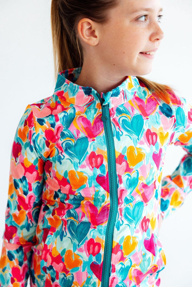 A radiant girl modeling the Heart Hustle Full-Zip Ruffle Girls Active Jacket in Turbo Teal, posing confidently to highlight the jacket's colorful design and functionality as a girls active jacket.