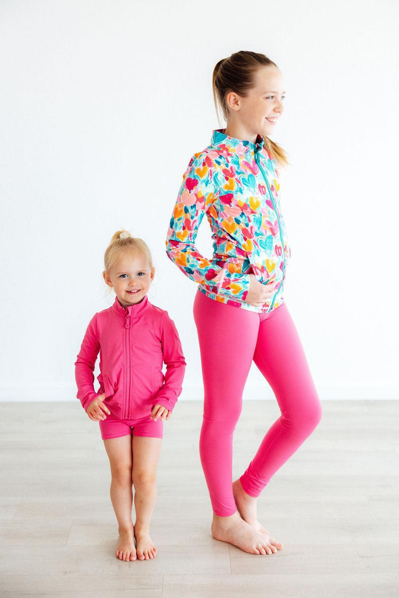 A young girl proudly posing next to her mother, both wearing colorful outfits with the Heart Hustle Full-Zip Ruffle Girls Active Jacket, showcasing their active lifestyle in trendy girls active jackets.