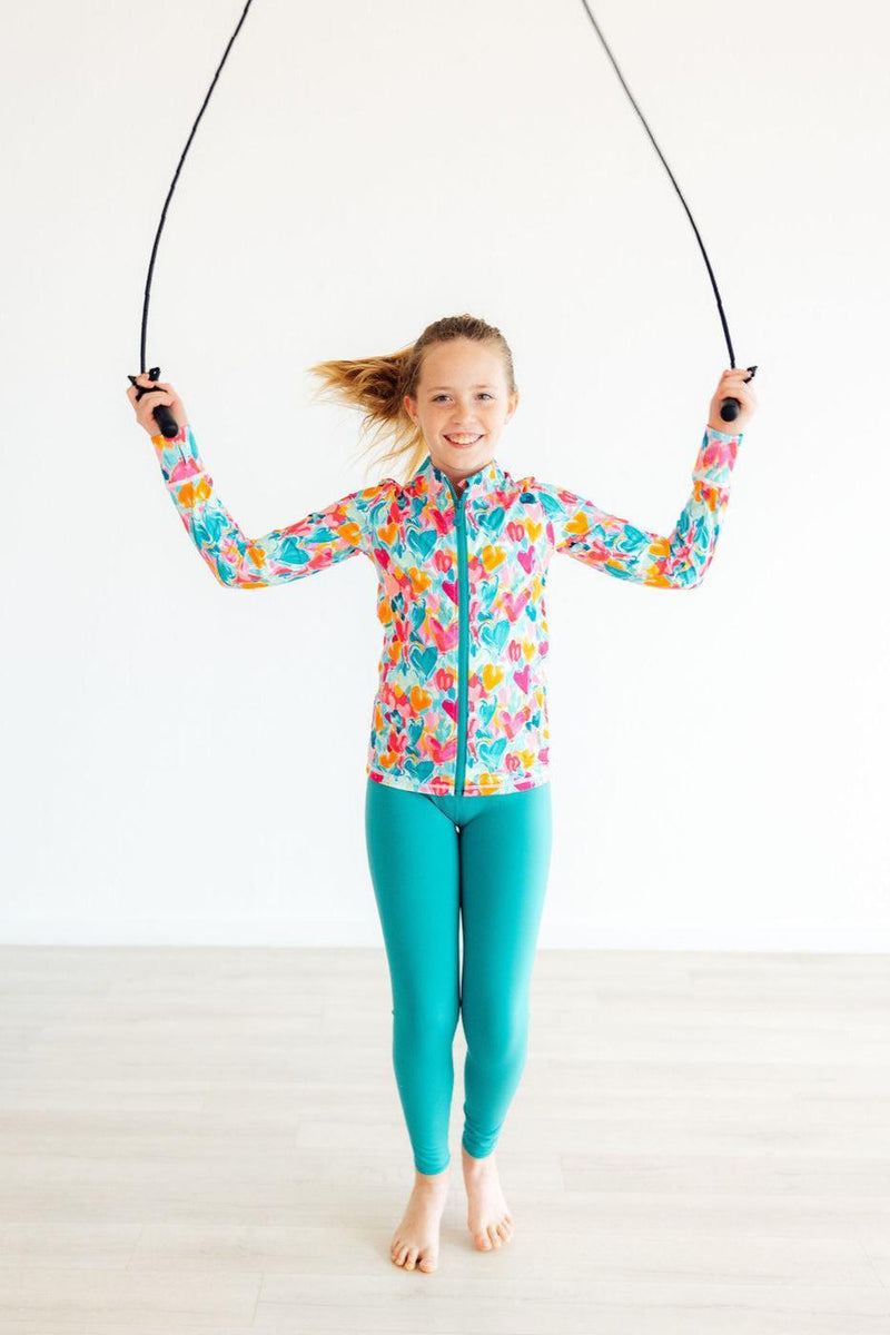 A cheerful girl wearing the Heart Hustle Full-Zip Ruffle Girls Active Jacket in vibrant Turbo Teal, jumping rope in an active pose. Perfect for active girls who love to move!