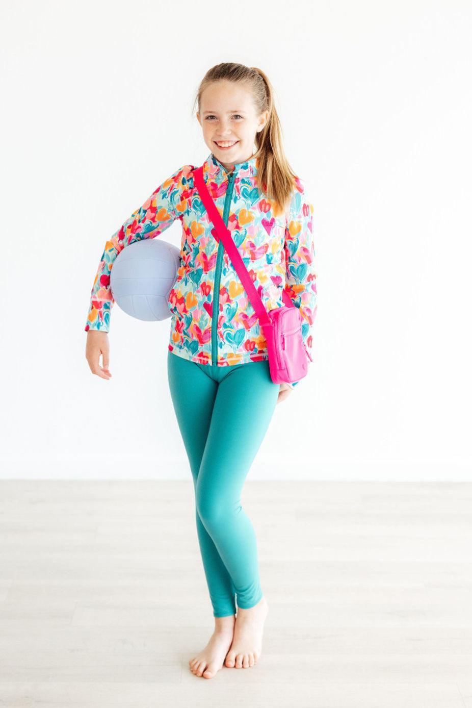 Smiling girl wearing Turbo Teal Active Leggings for Girls, colorful floral jacket, and holding a ball, showcasing stylish activewear perfect for young athletes.