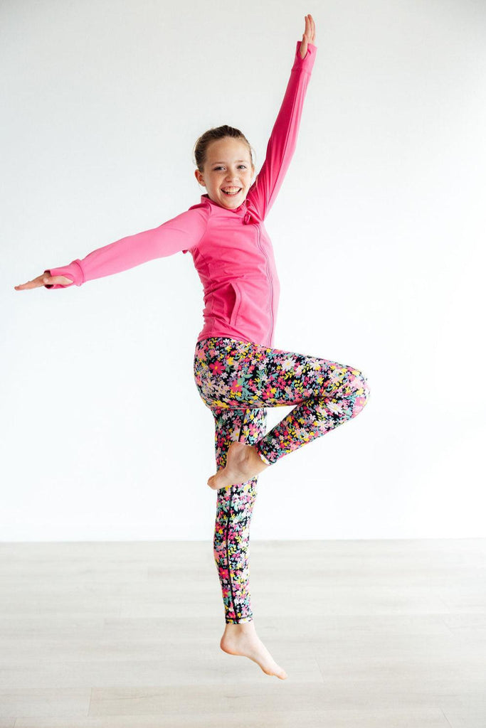A joyful girl wearing Flexible Floral Active Legging and a pink jacket, striking a playful pose on one leg in a bright and spacious environment.