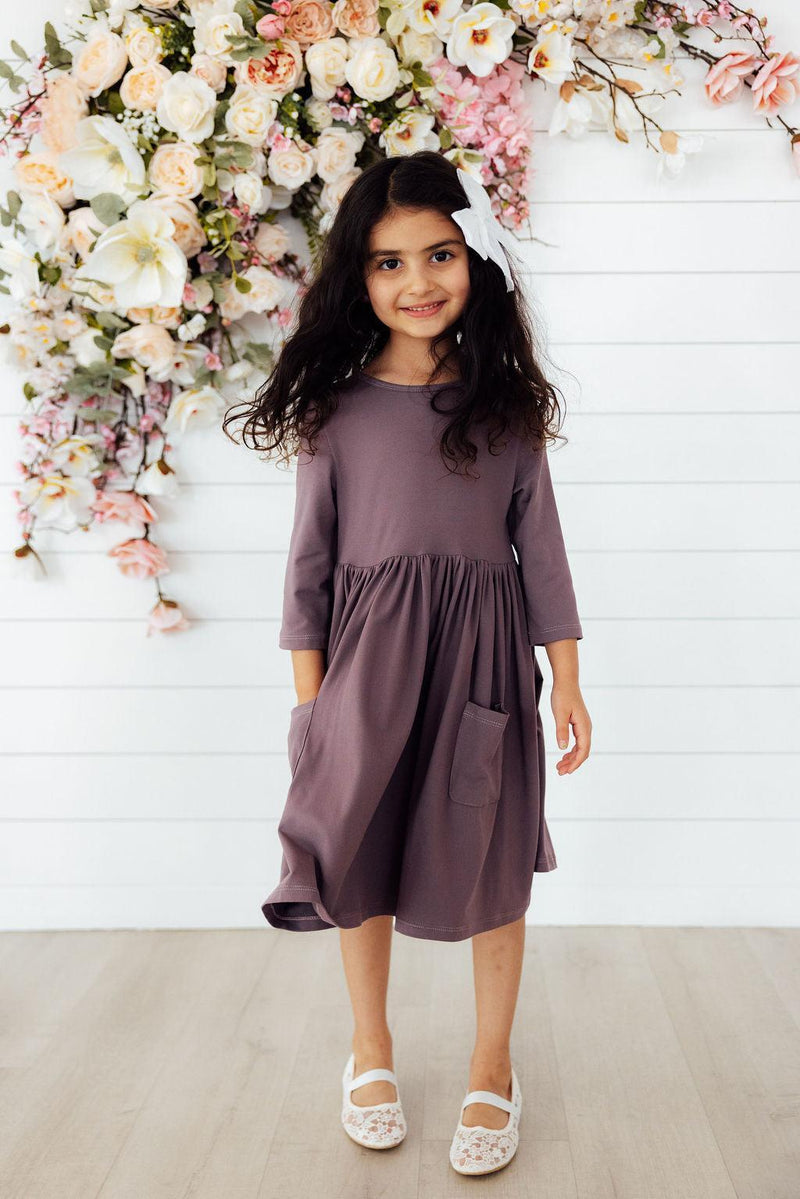 Smiling girl in a Vintage Violet 3/4 Sleeve Pocket Twirl Dress by Mila & Rose, displaying her twirl-friendly design and cute pockets against a floral backdrop.