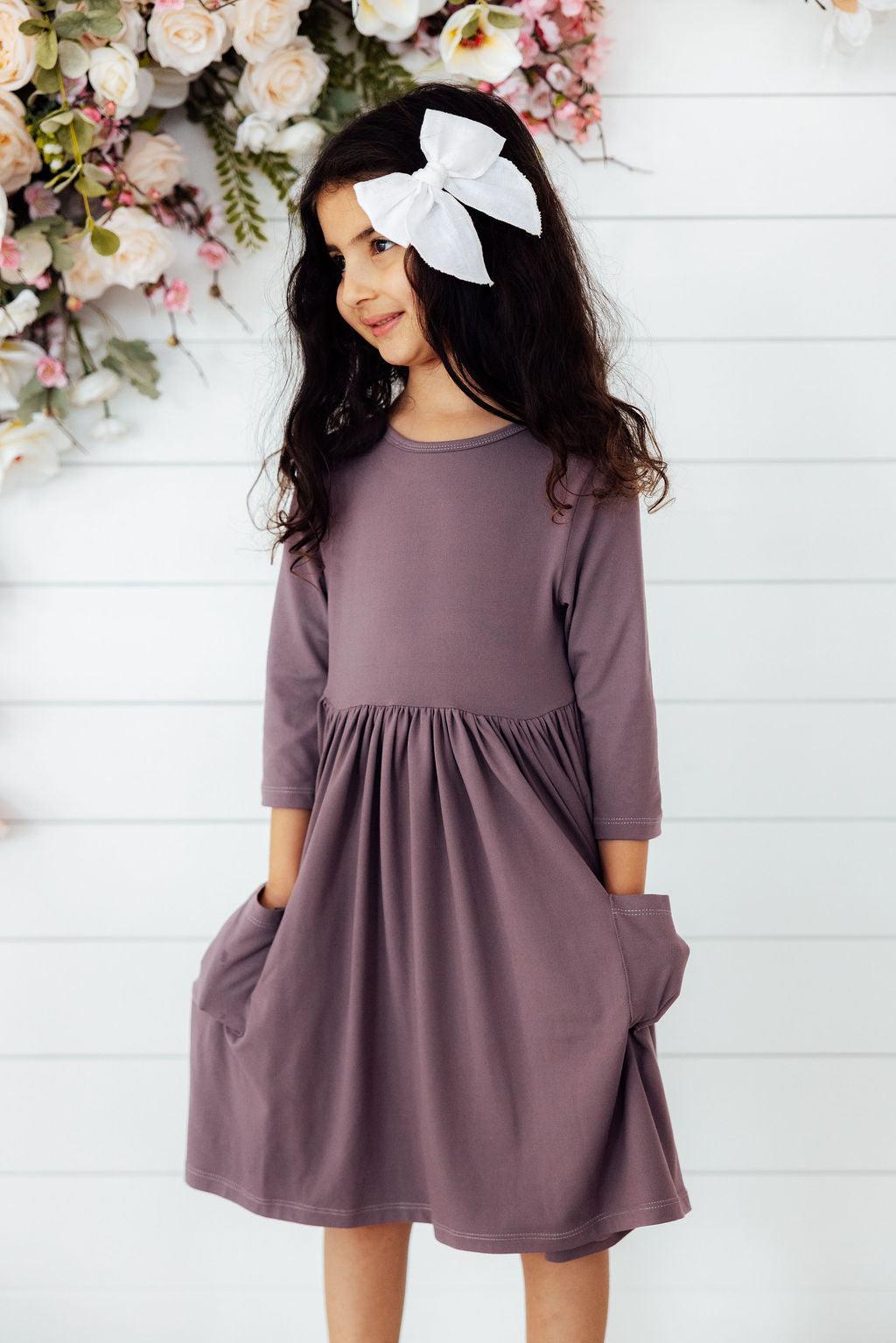 Girl wearing a Vintage Violet 3/4 Sleeve Pocket Twirl Dress by Mila & Rose, showcasing pockets and a playful twirl, surrounded by floral decorations.