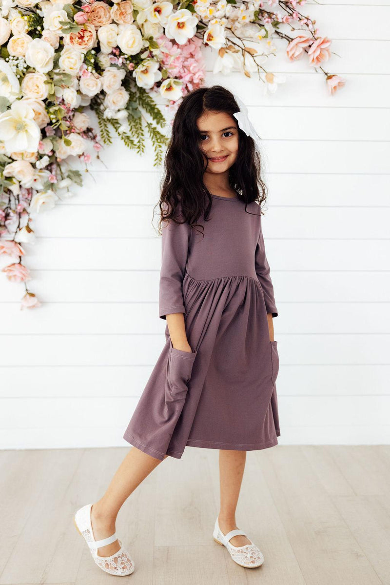A girl in a Vintage Violet 3/4 Sleeve Pocket Twirl Dress by Mila & Rose, smiling while standing in front of a floral backdrop.