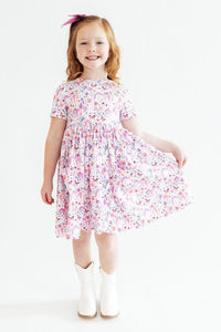 Young girl in Whimsical Woods Short Sleeve Pocket Twirl Dress with pink floral print, pink bow, and white boots.
