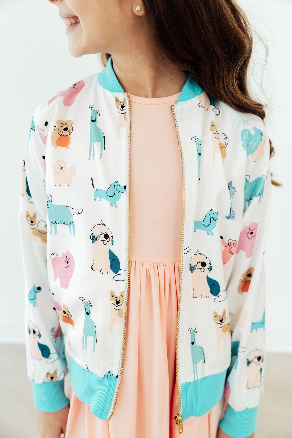 Young girl wearing Puppy Pals Satin Jacket with pastel dog prints and aqua cuffs over a peach dress.