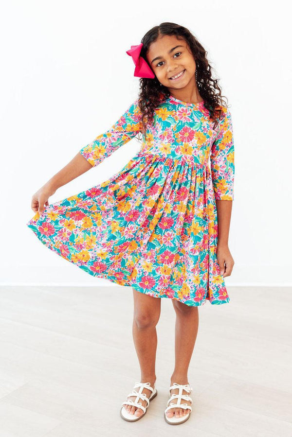 Floral Fiesta 3/4 Sleeve Pocket Twirl Dress worn by a smiling girl, twirling in a bright floral print with a pink bow.