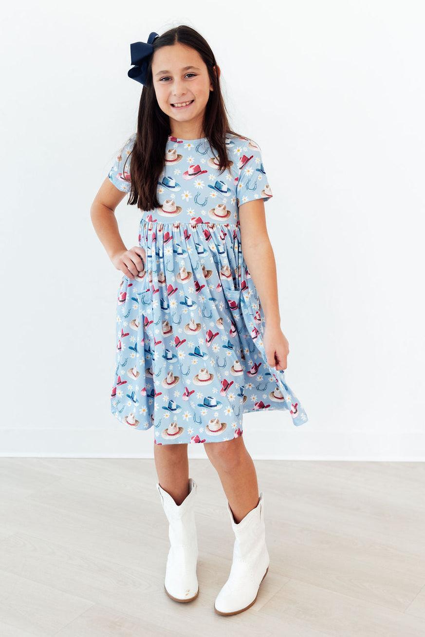 Happy girl wearing the Tip Your Hat Short Sleeve Pocket Twirl Dress For Girls, twirling in a park while highlighting its versatility and ease of movement.
