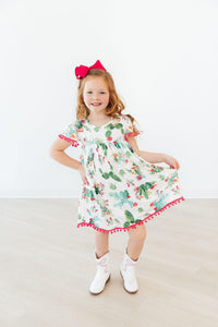 Smiling girl wearing Cute Cactus Pom Pom Dress with cactus print, pink pom-pom trim, and a red bow headband.