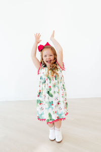 Little girl in Cute Cactus Pom Pom Dress with pink pom trim, red bow, arms raised.