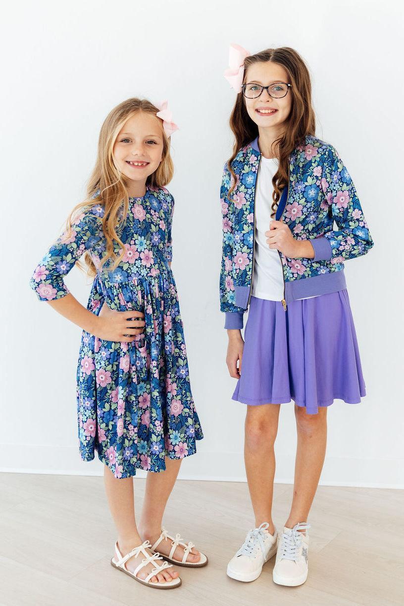 Two girls in blue floral outfits; one wears the Whimsy Satin Jacket, a blue satin bomber with lilac cuffs.