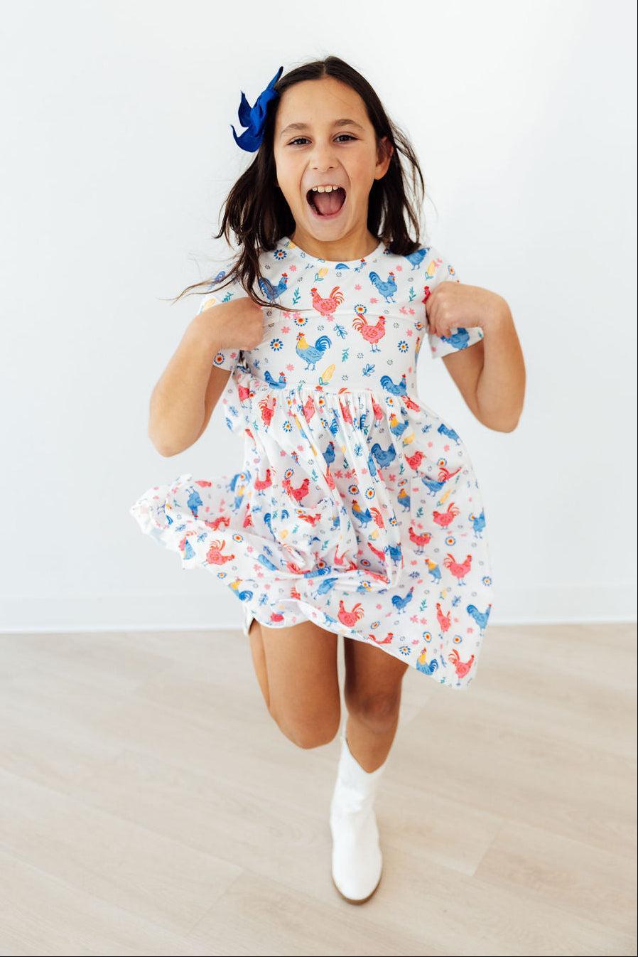 Full view of a child wearing the Rainbow Roosters Short Sleeve Pocket Twirl Dress For Girls, demonstrating its twirl silhouette and versatility as a pocket dress for girls.