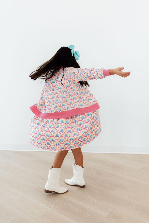 Child twirls in Watercolor Rainbows Satin Jacket with pastel rainbow print, pink cuffs and hem, blue hair bow, white boots.