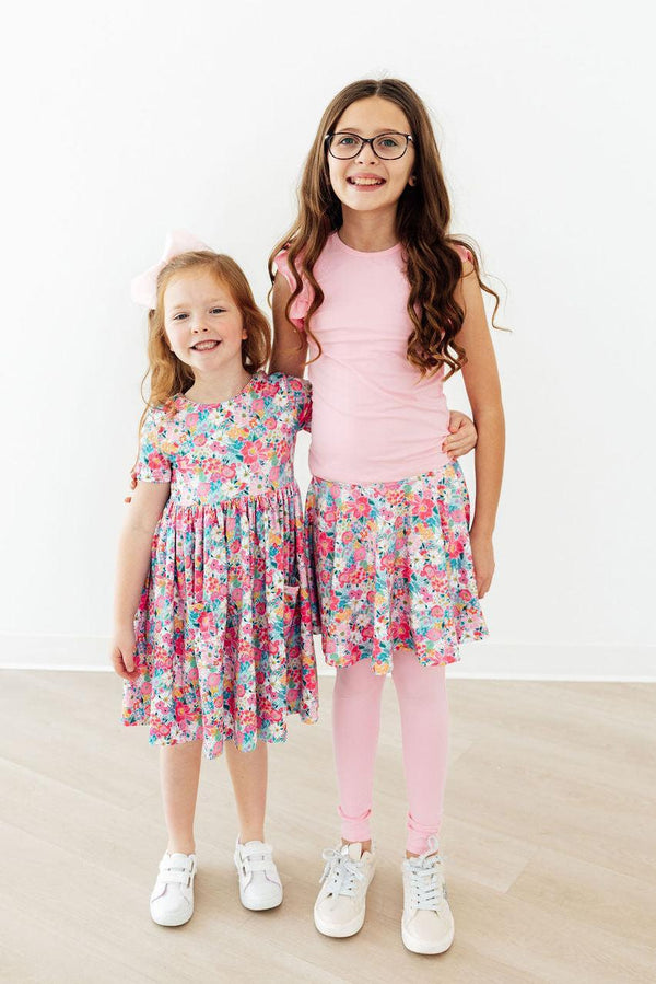 Two girls in floral twirl dresses pose together, one in a pink top and tights.