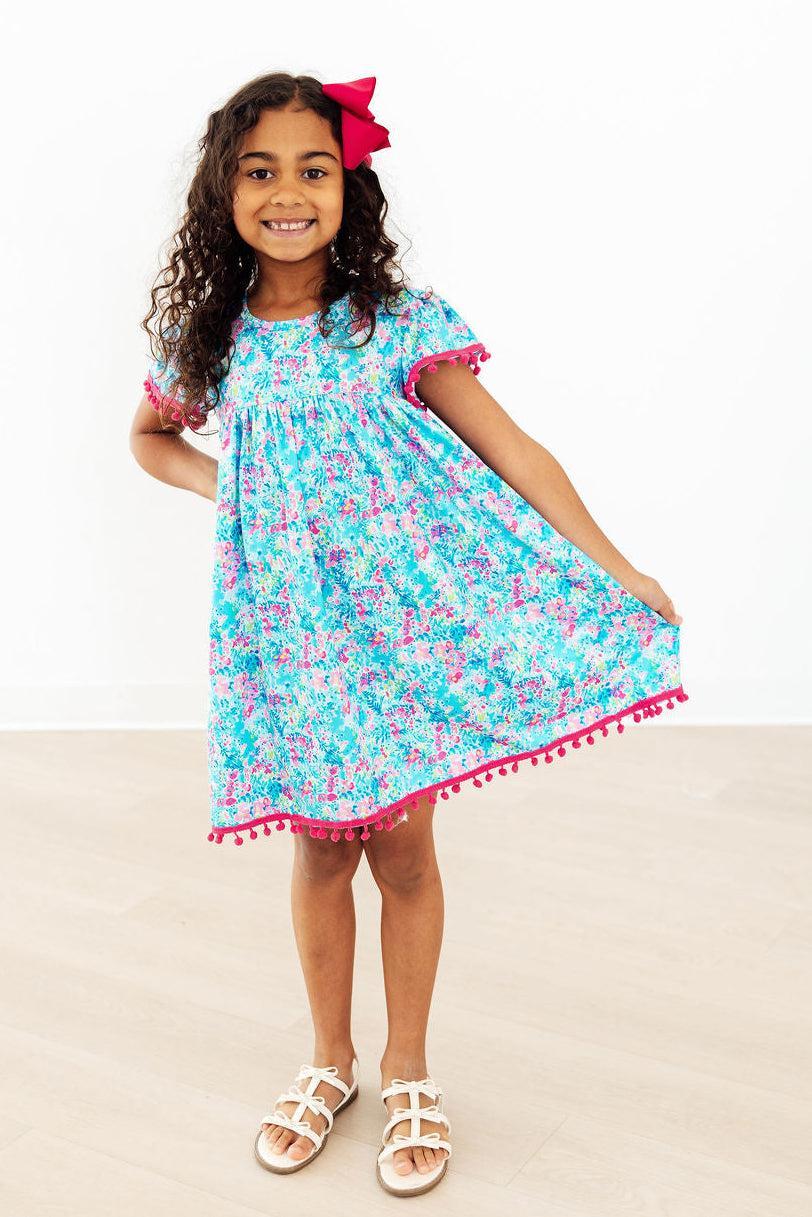 Aqua Garden Pom Pom Dress in blue floral print with pink pom-pom trim on sleeves and hem, worn by a girl.