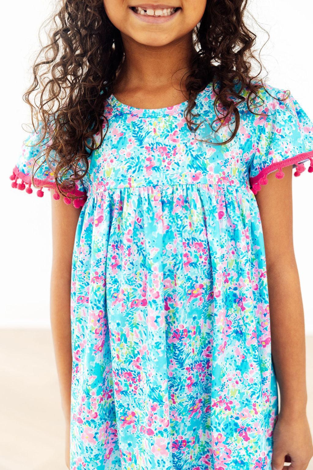 Aqua Garden Pom Pom Dress in blue floral print with pink pom pom trim on sleeves and hem.