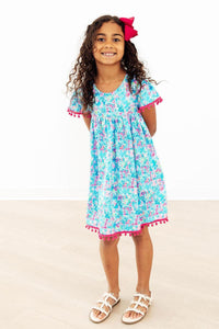 Aqua Garden Pom Pom Dress in blue floral print with pink pom pom trim on sleeves and hem worn by a smiling girl